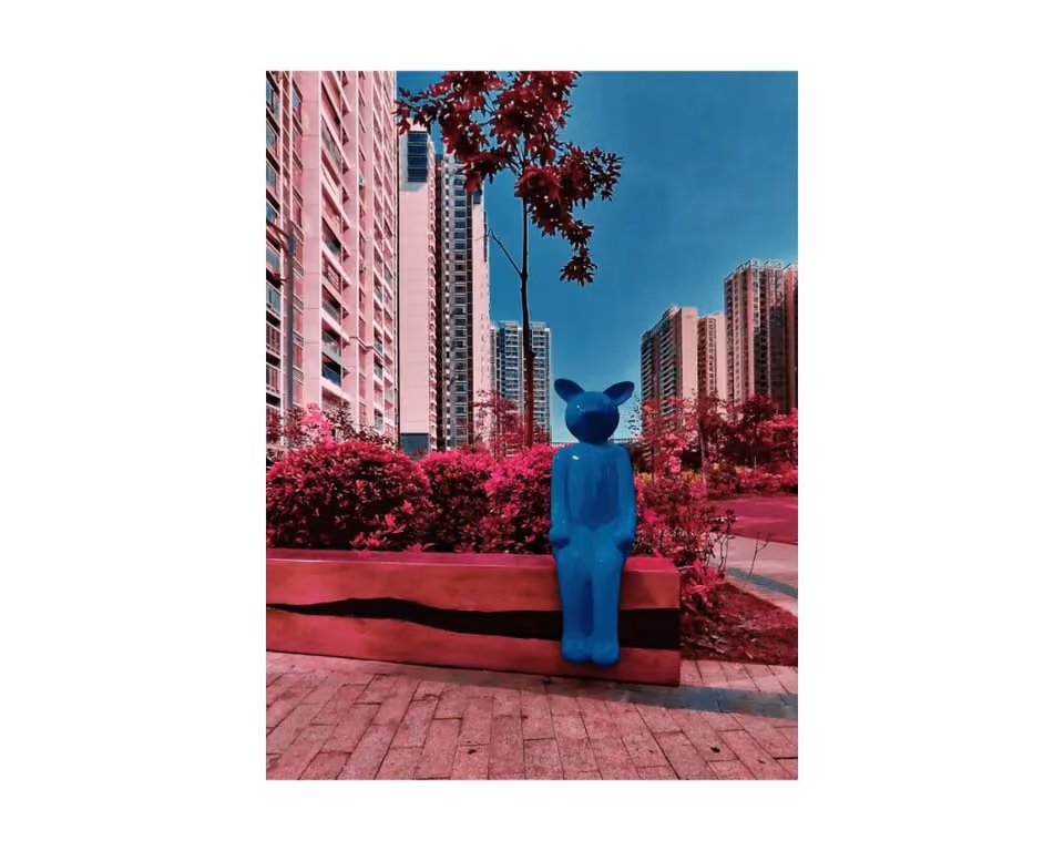 偏色画面中，蓝色人形雕塑坐在红色花坛上，背景是高层住宅楼